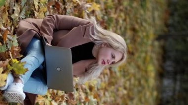 Dikey video. Çevrimiçi iletişim. Güzel kadın. Sonbahar doğası. Yaprakların üzerinde oturan gülümseyen kadın parkta dizüstü bilgisayar yazıyor..