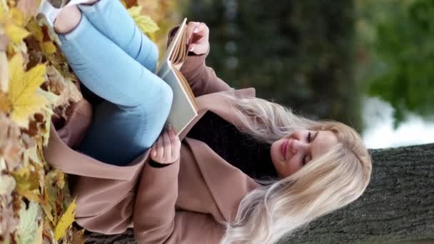 垂直录像 我喜欢看书 有灵感的女人美丽的秋天笑着的女士坐在公园里的地面斜树上 慢吞吞地笑着 — 图库视频影像