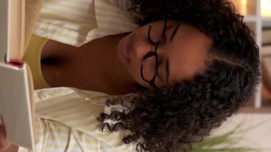 Dikey video. Okuma öğrencisi. Kitap dersi. Hafta sonu tatili. Çalışkan, neşeli kadın odadaki rahat halıda ödev öğreniyor..