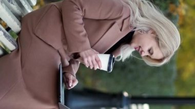 Dikey video. Serbest bir iş. Mutlu kadın. Sanal bağlantı. Park sokağında oturan gülümseyen kadın elinde bir fincan kahveyle dizüstü bilgisayarı yazıyor..