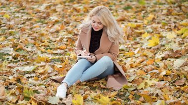 Romantik bir ruh hali. İlham verici bir kadın. Günlük anlar. Yaprakların üzerinde oturan mutlu kadın not defterine notlar yazıp sevinç duyuyor..