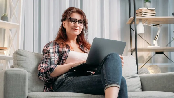 虚拟会议 成年妇女 家庭休闲 笑着坐在沙发上的中年女士 带着笔记本电脑在室内进行在线通话 — 图库照片
