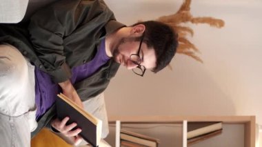 Dikey video. Ev ödevi. Boş zamanlarımda kitap okurum. Haftasonu dinlenme. Bardaklı barışçıl adam rahat oturma odasındaki rahat koltukta edebiyatın tadını çıkarıyor..