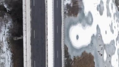 Dikey video. Drone karlı yolu. Hava trafiği. Asfaltlı buz köprüsü yüzeyi bulutlu kırsal bölgede donmuş göl..