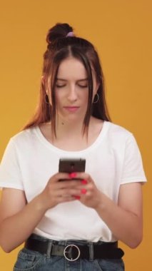 Dikey video. Akıllı telefon sorunu. Gadget sorunu. Portakal rengi arka planda izole edilmiş modern mobil cep telefonu cihazlı kızgın genç kız..