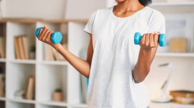 Dambıllar kalkıyor. Ev aerobiği. Spor elbiseli zayıf bir kadın. Ağırlık çalışıyor. Ev içi vücut geliştirme eğitimi alıyor..