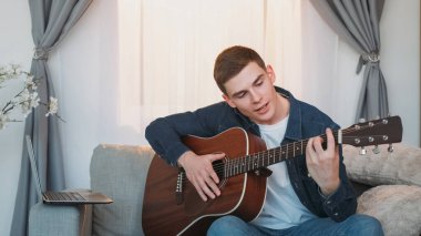 Çevrimiçi müzik dersi. Gitar dersi. Uzaktan eğitim. Evdeki kanepede dizüstü bilgisayar video konferansında telli çalgılar dersi veren adam..