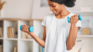 Dambıl egzersizleri. Aktif yaşam tarzı. Spor kıyafetleri içinde neşeli bir kadın. Evde ağırlık kaldırma antrenmanı yapıyor. Oturma odasında..