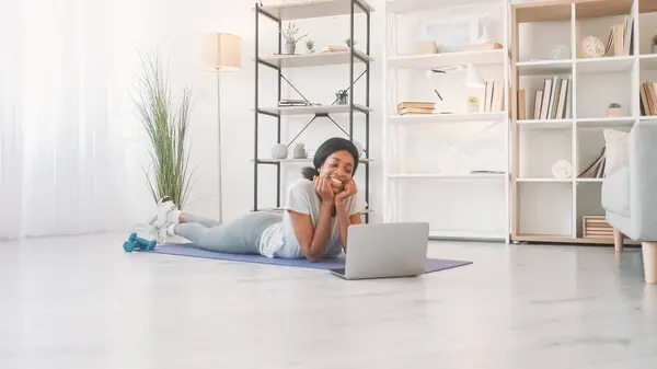 Online aerobics. Domestic training. Happy positive smiling woman enjoying internet fitness course at laptop video conference on sport mat at home in living room.