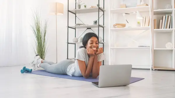 Online exercising. Home fitness. Cheerful young woman discussing aerobics training at laptop video meeting on yoga mat on floor in living room.