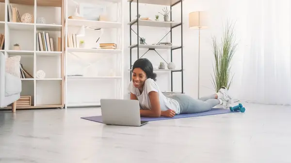 Video chat. Internet workout. Laptop communication. Happy woman talking at online call meeting on yoga mat after fitness training in home living room.