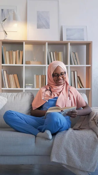 Doctor study. Medical education. Smiling woman student in hijab reading nurse course relaxing on sofa at hospital staff room.