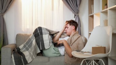 Video danışmanlığı. Uzaktan tıbbi yardım. Kötü baş ağrısı olan hasta bir adam dizüstü bilgisayarda konuşuyor. Evdeki oturma odasındaki kanepede tele-sağlık randevusu..