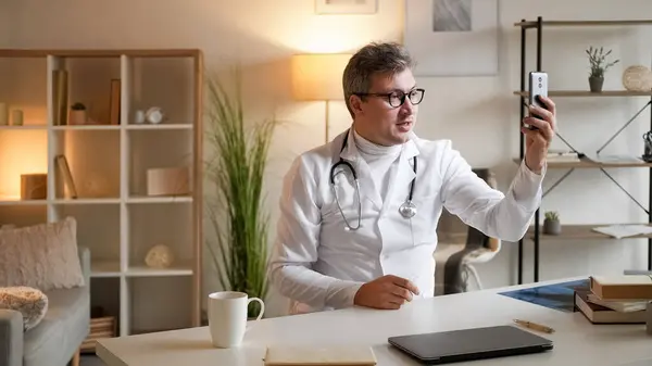 Medical video advice. Health consultation. Online meeting. Healthcare professional man talking at virtual conference on smartphone sitting at desk in clinic interior.