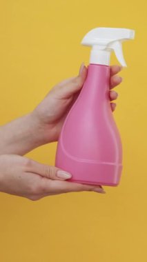 Dikey video. Temizlik spreyi. Ev hijyeni. Kadın elleri, sarı arka planda izole edilmiş yüzey deterjan sıvısıyla pembe şişe reklamı yapıyor..
