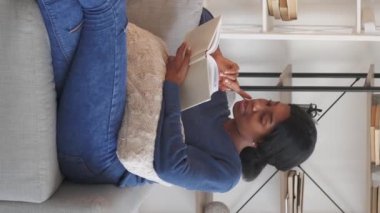 Dikey video. Edebiyat eğlencesi. Okuma hobisi. Kitap haftasonu. Neşeli genç bir kadın ilginç bir roman hikayesinin keyfini çıkarıyor. Evdeki rahat koltukta dinleniyor..