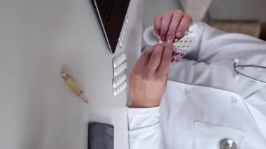 Dikey video. İlaç danışmanlığı. İnternetten sağlık randevusu. Beyaz üniformalı doktor, iş yerinde otururken dizüstü bilgisayardaki ilaç etkisini açıklıyor..