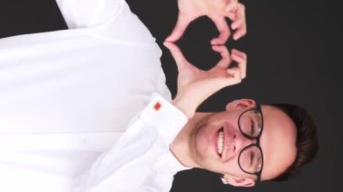 Dikey video. Aşk işareti. Romantik bir sevgi. Bağlılık bakımı. Gözlüklü neşeli adam siyah üzerine izole edilmiş kalp el hareketi gösteriyor..