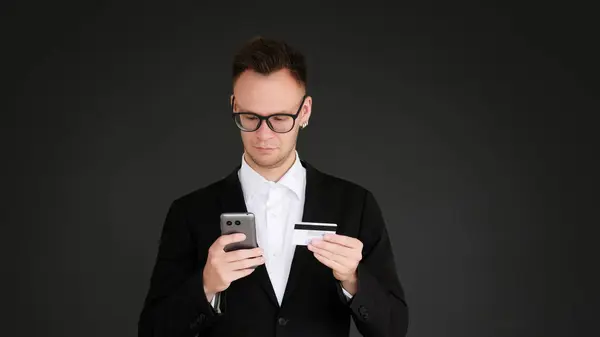 Mobile bank. Business transaction. Confident man in suit glasses using credit card phone isolated on dark black empty space background.