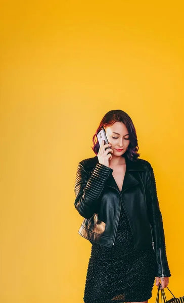 Talking phone. Mobile conversation. Serious concentrated woman in leather jacket speaking smartphone isolated on yellow copy space background.