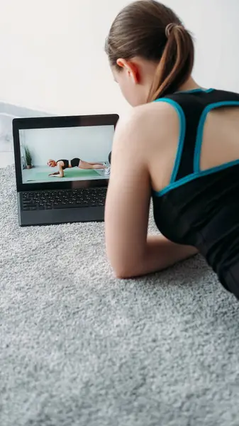 Virtual fitness class. Online training. Workout tutorial. Woman watching sport video program on laptop with athletic girl doing plank on screen at home.