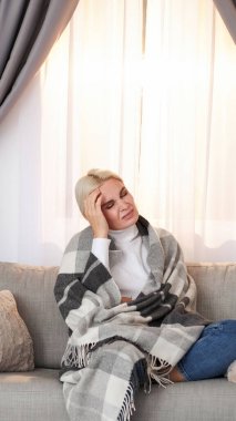 Kötü baş ağrısı. Soğuk acı. Yüksek sıcaklıkta bitkin düşmüş mutsuz hasta bir kadın. Evdeki fotokopi odasındaki kanepede dinlenen battaniyeye sarılı acı veren semptomlar..