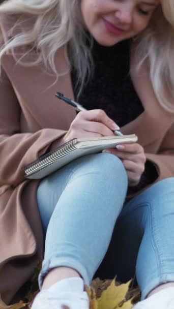 バーティカルビデオ ロマンチックな手紙 インスパイアされた女性 秋の美しさ 葉の上に座っている笑顔の女性はノートにノートを作ります — ストック動画