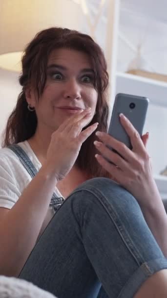 バーティカルビデオ モバイル通信 興奮する女性 バーチャルライフ ライトルームのインテリアでスマートフォンをスクロールする床に座っている中年の幸せな女性 — ストック動画