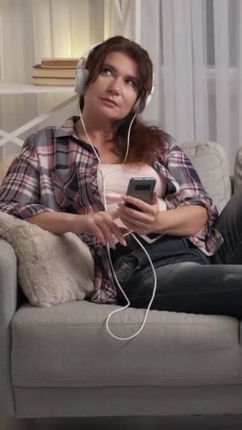 バーティカルビデオ 音楽の喜び リラックスした大人女性 ホームレジャー スマートフォンでスマートフォンとソファーに座ってヘッドフォンで笑顔のカジュアルな女性 — ストック動画