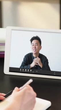 Dikey video. Online danışmanlık. İş koçluğu. Kadın öğrenci, iş yerindeki sanal toplantıda uzman bir adamın tablet ekranda konuşması üzerine notlar yazarak teslim ediyor..