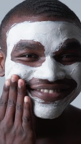 バーティカルビデオ 人間の化粧品 フェイシャルマスク グレーの背景で笑顔の白い粘土をきれいにする顔の皮の気孔を適用することを楽しんでいる確信した満足した男 — ストック動画