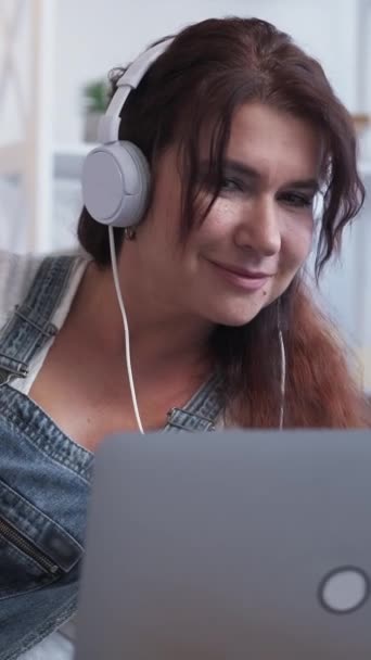 バーティカルビデオ オーディオ技術 インスパイアされた女性 ホームリラックス ライトルームのインテリアでラップトップで音楽を聞くヘッドフォンのハッピー中年の女性 — ストック動画