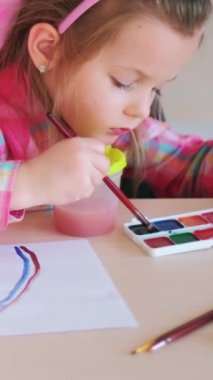 Dikey video. Çocuk ressam. Yaratıcı bir eğlence. Yetenekli çocuklar için okuldaki masada suluboya ve boya fırçasıyla gökkuşağı resmi yapan küçük bir kız..