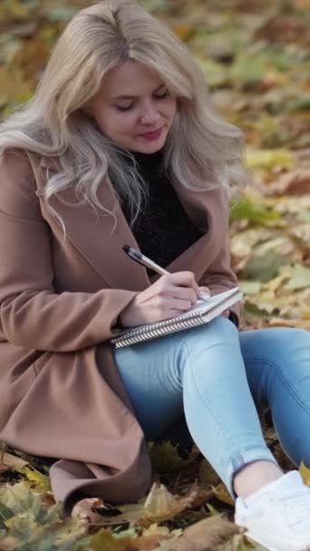 バーティカルビデオ 手紙を書いている ロマンチックな女性 秋のインスピレーション 葉の上に座っている笑顔の女性は公園のノートにノートを作ります — ストック動画