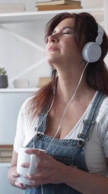 Dikey video. Müzik aşığı. Tatmin olmuş bir kadın. Evde rahatla. Orta yaşlı, kulaklıklı, koltukta oturan, aydınlık odada kahve içen bir kadından esinlendim..
