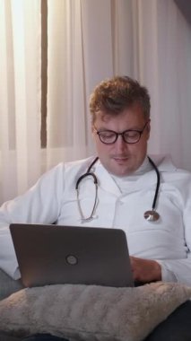 Dikey video. Boş zamanlarında çalış. Doktor molası. Çevrimiçi iletişim. Rahatlamış sağlık uzmanı, beyaz üniformalı, dizüstü bilgisayarla koltukta oturuyor..