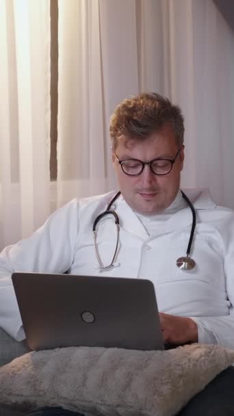 垂直录像 工作休闲 博士休息 在线交流 宽松的医疗保健专家 身穿白色制服 坐在轻便的沙发上 在笔记本电脑上打字 — 图库视频影像