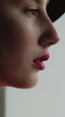 Dikey video. Profesyonel makyaj. Güzellik trendi. Sulu kırmızı rujlu, şık bir kadın mükemmel yüz derisi altın küpeler gri arka planda siyah şapkalar.