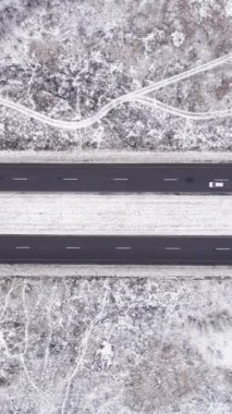 Dikey video. Hava karlı görünüyor. Kırsal kış yolu. Beyaz donmuş tarlalar, asfalt pist ve geçen arabalarla trafik yolu bulutlu hava aracı..