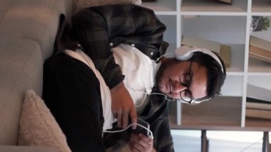 Dikey video. Mobil müzik. Eğlenceli bir şarkı. Çalma listesi eğlencesi. Mutlu adam telefonda kulaklıkla en sevdiği melodiyi dinliyor. Oturma odasındaki kanepede dinleniyor..