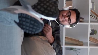Dikey video. Müzik zevki. Ses aygıtı. Çalma listesi eğlencesi. Ev tatili. Mutlu gülümseyen adam telefonda en sevdiği şarkıyı dinliyor koltukta, rahat oturma odasında..