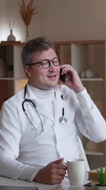 Dikey video. Doktor telefon konuşması. Cep telefonu. İş molası. Hücre iletişiminin keyfini çıkaran pozitif hastane uzmanı klinik masasında oturup kahve içiyor..