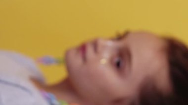 Dikey video. Neşeli çocuk. Sevimli çocuk. Yüzünde sahte parlak çillerle gülümseyen sevimli küçük bir kız. Renkli küpeler, sarı arka planda 2 'lik izole bir şekilde belirdi..