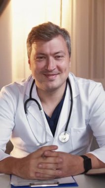 Dikey video. Doktor konsültasyonu. Tıbbi destek. Tele-sağlık yardımı. Beyaz hastane üniformalı olumlu bir adam online randevusunda ışık odasında masasında oturuyor..