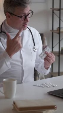 Dikey video. İlaç reçetesi. Çevrimiçi doktor danışmanlığı. Laptop konferansında klinikteki işyerinde oturup hap tedavisi hakkında konuşan profesyonel bir doktor..