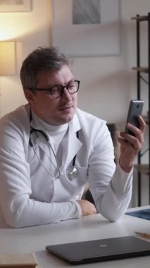 Dikey video. Tıbbi uzaktan tavsiye. Sağlık danışmanlığı. Çevrimiçi toplantı. Cep telefonu konferansında konuşan sağlık uzmanı kliniğin içindeki masada oturuyor..