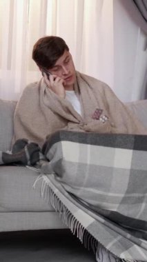 Dikey video. Gezici danışmanlık. Grip tedavisi. Ateşli, ekoseli hasta bir adam akıllı telefonuyla evdeki oturma odasındaki kanepede oturan haplardan bahsediyor..