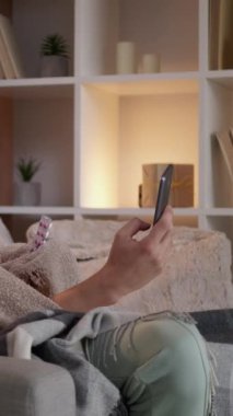 Dikey video. Çevrimiçi sağlık hizmeti. Reçeteli ilaçlar. Battaniyeye sarılı yorgun bir adam akıllı telefon konuşmasında evdeki kanepede oturup ilaçla ilgili konuşuyor..