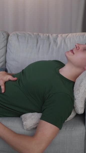 バーティカルビデオ ベリー痛み スタマ感染症 自宅にソファーを置くウイルスの症状に苦しんでいる感じの腹部の手の疲れた男 — ストック動画