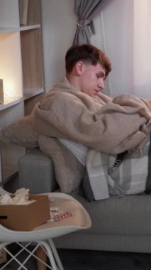 Dikey video. Grip belirtileri. Ateş baş ağrısı. Mevsimsel enfeksiyon. Hasta adam battaniyeye sarılı olarak kanepede oturuyor ve evde ilaçla soğuk algınlığı yüzünden öksürüyor..